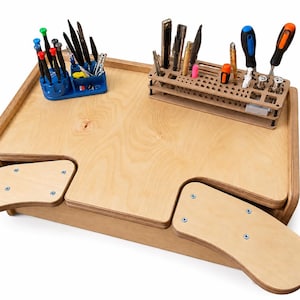 May include: A light-colored wooden workbench with two extended armrests, featuring a blue plastic tool organizer filled with various screwdrivers and a rectangular wooden tool rack holding multiple metal tools and screwdrivers.