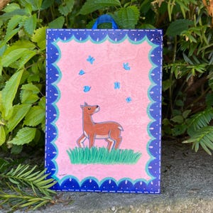 May include: A hand-painted wooden sign with a pink background and a blue border. The sign features a brown deer with a white face and a green grassy area at the bottom. There are blue butterflies flying around the deer.