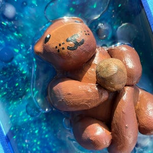 May include: A brown ceramic figurine of a bear holding a round object in its paws. The bear is sitting in a blue liquid with glitter.