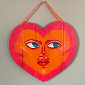 Puede incluir: Un letrero de madera en forma de corazón pintado en tonos de rosa y naranja con una cara estilizada. La cara tiene ojos azules, una boca roja y una nariz.