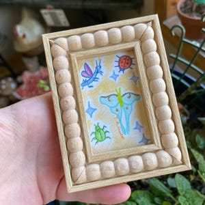 Puede incluir: Un pequeño marco rectangular de madera con un acabado claro. El marco rodea una colorida ilustración de insectos y una mariposa. La obra presenta una mariposa azul, una mariquita, una libélula y un escarabajo verde. La obra está firmada.