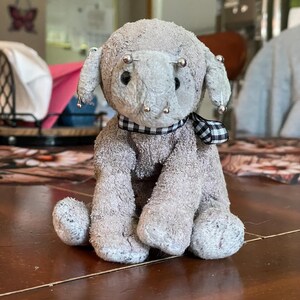 May include: A gray plush elephant toy with a textured fabric surface, featuring black button eyes and several silver-toned sewing pins inserted into its head and ears. A black and white checkered ribbon is tied around its neck.