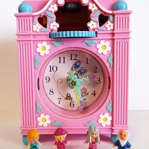 Puede incluir: Un reloj de juguete rosa con una esfera redonda con un diseño de mariposa y abeja. El reloj está decorado con flores blancas y detalles azules. Cuatro pequeñas figuras están de pie frente al reloj.