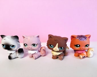 Puede incluir: Cuatro figuras de gatos de colores con ojos grandes y detalles. Los gatos son de varios colores, incluyendo blanco y negro, rosa, marrón y naranja, y están posando sentados. El fondo es rosa liso.