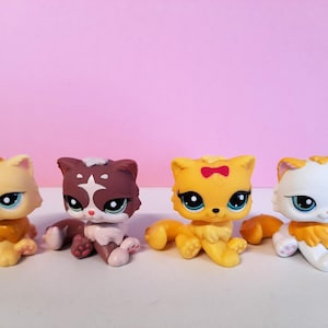 Könnte beinhalten: Vier farbenfrohe Katzenfiguren in Gelb-, Braun- und Weißtönen, jede mit großen blauen Augen. Eine Katze hat eine rote Schleife. Die Spielzeuge sitzen auf einer weißen Oberfläche vor einem rosa Hintergrund.