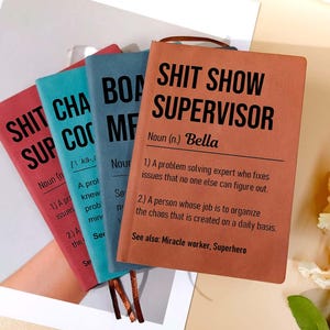 Może przedstawiać: Stos czterech notatników z zabawnymi tytułami. Notatniki są w odcieniach czerwieni, turkusu i niebieskiego, z przednim notatnikiem w czerwonobrązowym kolorze. Tekst na okładkach notatników brzmi "SHIT SHOW SUPERVISOR" i inne podobne zwroty.