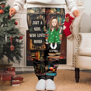 Może przedstawiać: Czarne spodnie od piżamy z motywem półki na książki oraz napisem "JUST A GIRL WHO LOVES BOOKS" i imieniem "SOPHIA". Spodnie przedstawiają dziewczynkę z kreskówki w zielonej sukience trzymającą książkę. Osoba ma na sobie białe kapcie.