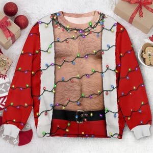 Op de afbeelding: Een sweatshirt met lange mouwen en ronde hals met een print van een behaarde borst, een zwarte riem met een gouden gesp en witte randen, alles omwikkeld met een snoer van kleurrijke kerstverlichting tegen een felrode achtergrond.
