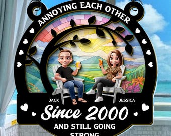May include: A black decorative sign with the text "ANNOYING EACH OTHER" and "Since 2000 AND STILL GOING STRONG." It features a couple sitting in chairs, holding drinks, with a colorful stained-glass background.