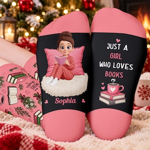 May include: A pair of socks with a pink and black colour scheme. One sock features a cartoon character reading a book on a white rug with the name Sophia printed below. The other sock displays the text JUST A GIRL WHO LOVES BOOKS above a coffee cup.