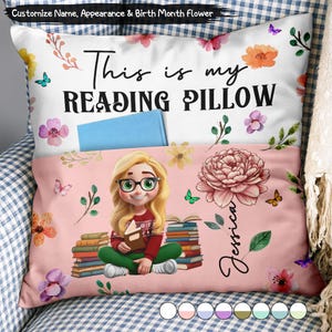May include: A white and pink throw pillow with a pocket containing a blue book. The text reads This is my READING PILLOW. It features a cartoon illustration of a blonde person with glasses sitting by a stack of books, a large flower, and the name Jessica.