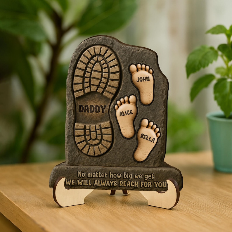 May include: A wooden plaque with a footprint and the words "Daddy", "Alice", "John", and "Bella" engraved on it. The plaque also has the text "No matter how big we get, we will always reach for you".