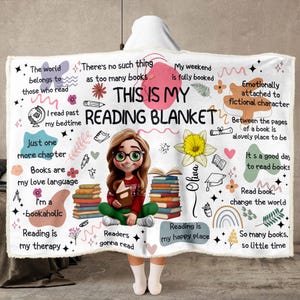 Può includere: Una coperta con cappuccio bianca con un'illustrazione centrale di una persona con gli occhiali che legge un libro, circondata da pile di libri, un fiore giallo e testo tra cui "THIS IS MY READING BLANKET" e varie citazioni sui libri.