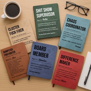 May include: A collection of notebooks with humorous titles like "Cluster Fuck Fixer" and "Chaos Coordinator." Each notebook has a different color cover and includes a definition. A cup of coffee and a pair of glasses are also in the image.