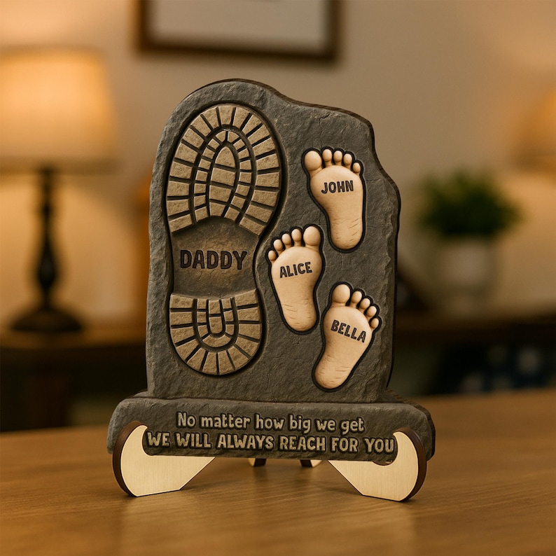 May include: A brown wooden plaque with a footprint design. The plaque has the words "Daddy", "Alice", "John", and "Bella" engraved on it. The plaque also has the text "No matter how big we get WE WILL ALWAYS REACH FOR YOU".