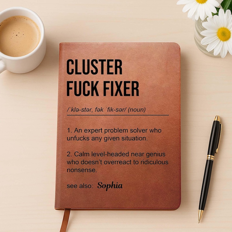 May include: A brown notebook with the text "CLUSTER FUCK FIXER" in bold black letters. The notebook includes a definition and the name "Sophia". A black pen and a cup of coffee are next to the notebook.