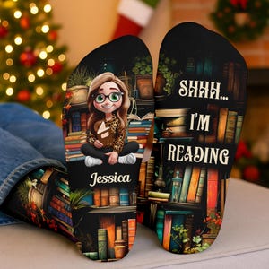 May include: Black socks featuring a cartoon illustration of a girl reading, surrounded by books. The socks display the text "SHHH... I'M READING" and the name "Jessica". A comfortable and stylish accessory for book enthusiasts.