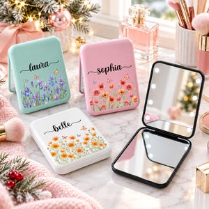 May include: Three rectangular compact mirrors with LED lights, featuring floral prints and the names laura, sophia, and belle. One mirror is open, showing internal lights, while two are closed. The cases are mint green, pink, and white.