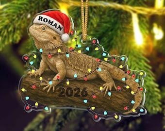 May include: A flat, clear acrylic Christmas ornament featuring a bearded dragon resting on a brown log. The lizard wears a red and white Santa hat with the name ROMAN printed on it. The log displays the year 2026 and is wrapped in colorful lights.