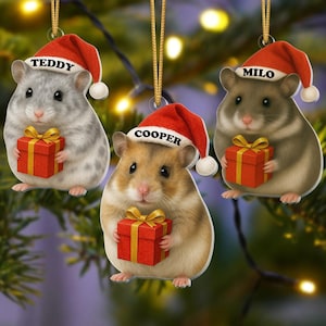 Könnte beinhalten: Drei flache Hängedekorationen in Form von Hamstern, die jeweils eine rote Weihnachtsmütze mit weißem Bommel tragen. Die Hamster sind grau, beige und braun und halten jeweils ein rotes Geschenk mit goldener Schleife. Die Mützen sind mit den Namen Teddy, Cooper und Milo personalisiert.