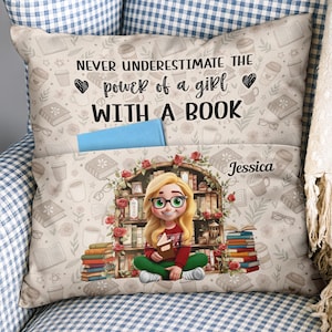 Puede incluir: Un cojín decorativo con un diseño temático de libros. El cojín presenta el texto "Never underestimate the power of a girl with a book". Una chica de dibujos animados con gafas y un libro está en el centro. El cojín tiene un bolsillo con un libro azul.