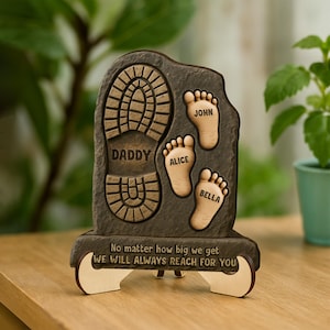 May include: A wooden plaque with a footprint and the words "Daddy", "Alice", "John", and "Bella" engraved on it. The plaque also has the text "No matter how big we get, we will always reach for you".
