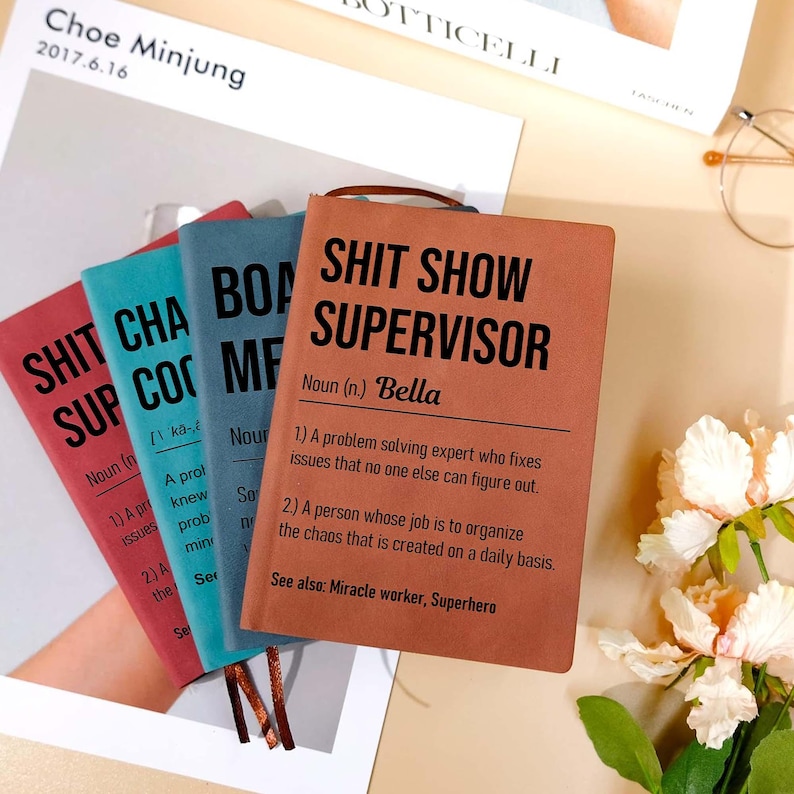 May include: A stack of four notebooks with humorous titles. The notebooks are in shades of red, teal, and blue, with the front-most notebook in a reddish-brown color. The text on the front of the notebooks reads "SHIT SHOW SUPERVISOR" and other similar phrases.