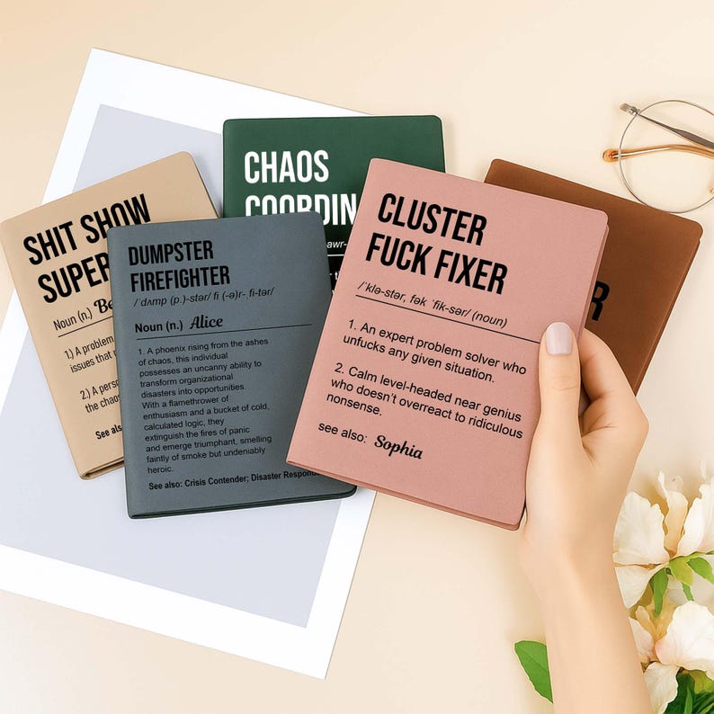 May include: A collection of notebooks with humorous definitions. The notebooks are in various colors, including tan, gray, green, and pink. One notebook reads "Cluster Fuck Fixer," and another says "Dumpster Firefighter."