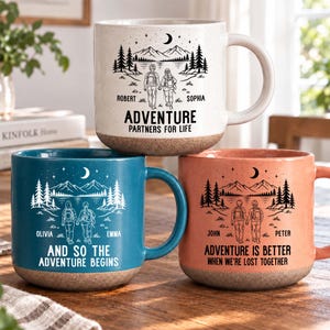 May include: Three ceramic mugs in cream, blue, and peach, each featuring a speckled finish and a natural-colored base. The mugs display black line art of two people hiking toward mountains under a crescent moon, with personalized names and text.