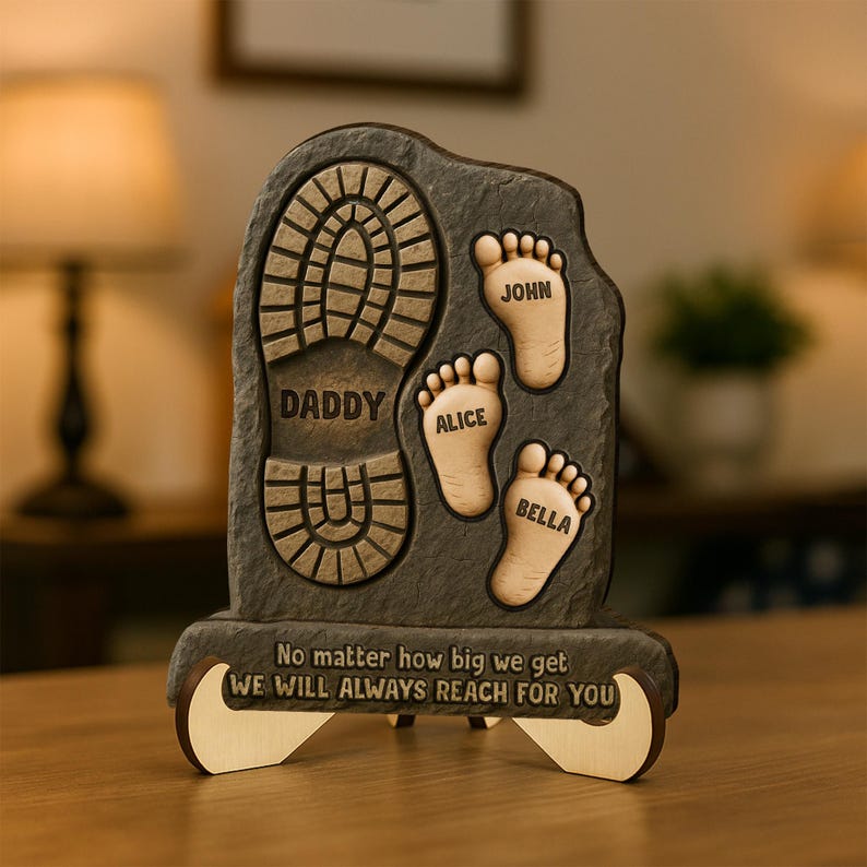 May include: A brown wooden plaque with a footprint design. The plaque has the words "Daddy", "Alice", "John", and "Bella" engraved on it. The plaque also has the text "No matter how big we get WE WILL ALWAYS REACH FOR YOU".