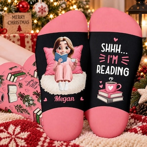 Può includere: Un paio di calzini rosa e neri con un personaggio dei cartoni animati che legge un libro su un tappeto bianco con il nome Megan. L'altro calzino mostra il testo SHHH... I'M READING sopra una pila di libri e una tazza da caffè rosa.