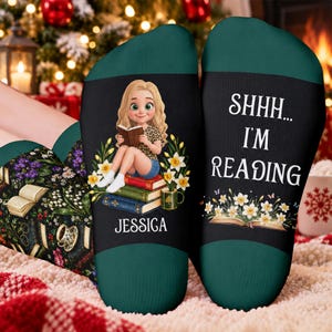 Könnte beinhalten: Ein Paar dunkelgrüne Socken mit einer Cartoon-Illustration einer blonden Person, die auf einem Bücherstapel sitzt und liest, umgeben von gelben Blumen. Der Text lautet SHHH... I'M READING und der Name JESSICA steht darunter.