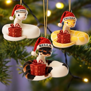 May include: Three hanging Christmas ornaments featuring snakes in red Santa hats. One white snake labeled Buddy, one yellow snake labeled Cherry, and one black, white, and brown snake labeled Bella. Each snake holds a red gift box with gold stars.