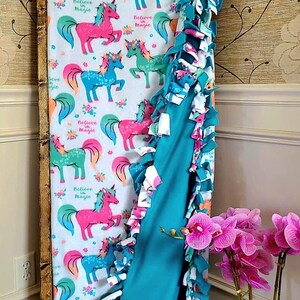 Könnte beinhalten: Eine türkisblaue und weiße Batik-Fleece-Decke mit einem Einhornmuster und dem Text "Believe in Magic". Die Decke ist gefaltet und auf einem Holzrahmen ausgestellt.