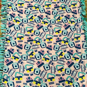 May include: A pink fleece tie blanket with a repeating pattern of vintage cameras. The cameras are in various colors, including yellow, blue, pink, and green. The text "Glamour Shot", "Snap!" , "Click", "Smile", and "Flash" appears on the blanket.