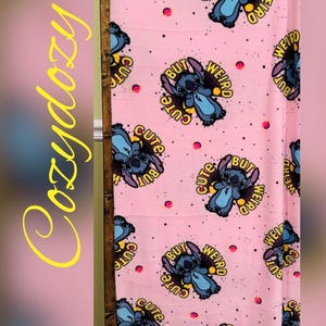 Pu&ograve; includere: Coperta rosa con un motivo ripetuto di un personaggio dei cartoni animati blu e la scritta "Cute But Weird". La coperta ha uno sfondo rosa con piccoli puntini rosa. La parola "Cozydozy" &egrave; scritta in giallo sul lato.