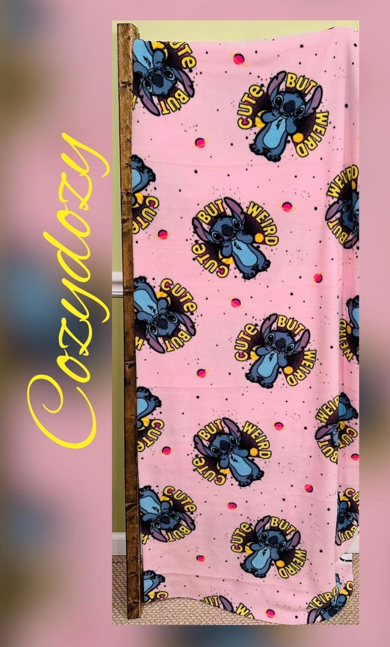 Pu&ograve; includere: Coperta rosa con un motivo ripetuto di un personaggio dei cartoni animati blu e la scritta "Cute But Weird". La coperta ha uno sfondo rosa con piccoli puntini rosa. La parola "Cozydozy" &egrave; scritta in giallo sul lato.