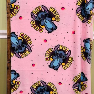 Pu&ograve; includere: Coperta in pile rosa con un motivo ripetuto di un personaggio dei cartoni animati blu e la scritta "Cute But Weird" in giallo e nero. Il design include pianeti rosa e punti neri.