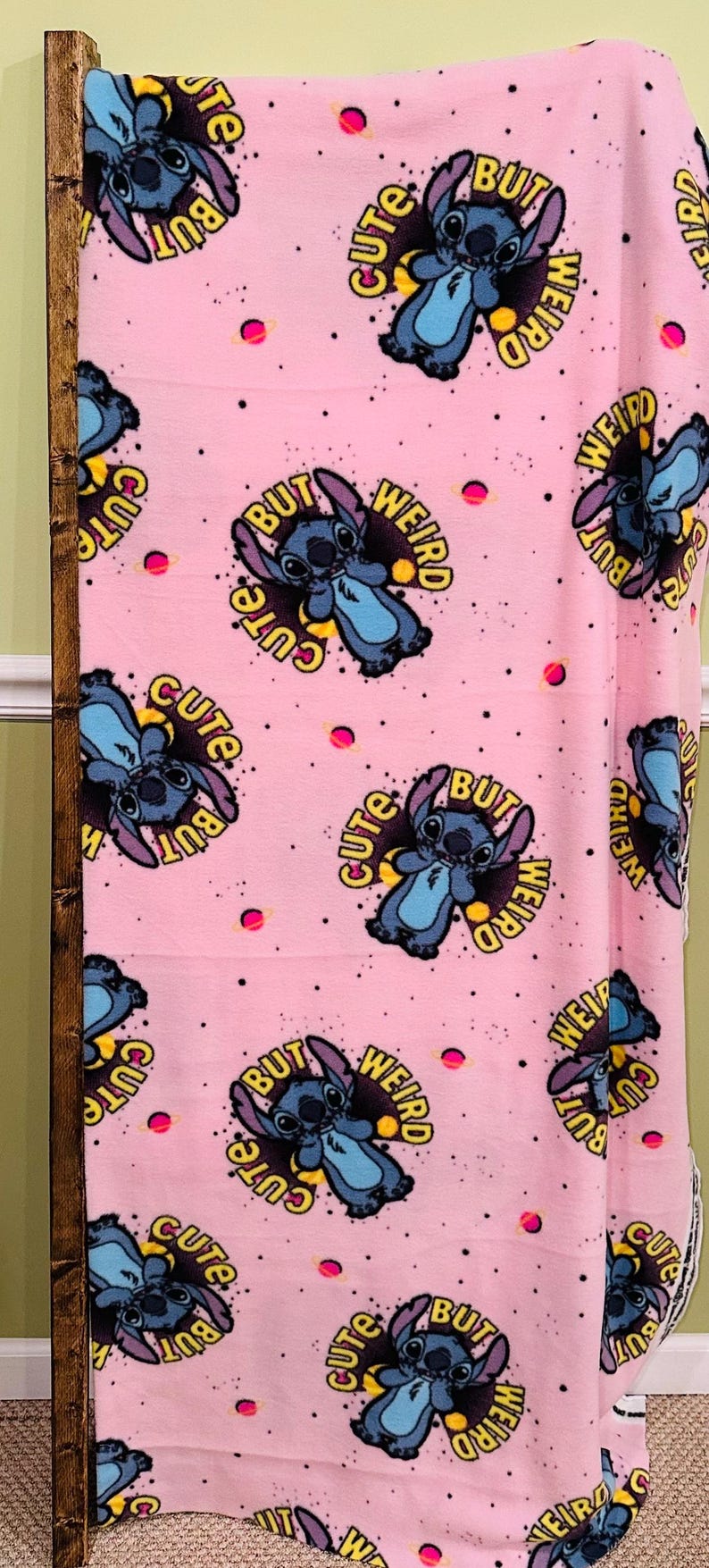 Pu&ograve; includere: Coperta in pile rosa con un motivo ripetuto di un personaggio dei cartoni animati blu e la scritta "Cute But Weird" in giallo e nero. Il design include pianeti rosa e punti neri.