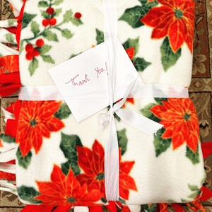 K&ouml;nnte beinhalten: Eine wei&szlig;e Fleece-Decke mit einem roten Weihnachtsstern-Druck und gr&uuml;nen Bl&auml;ttern. Die Decke ist mit einem wei&szlig;en Band gebunden und hat einen handgeschriebenen Zettel mit der Aufschrift "Danke!"