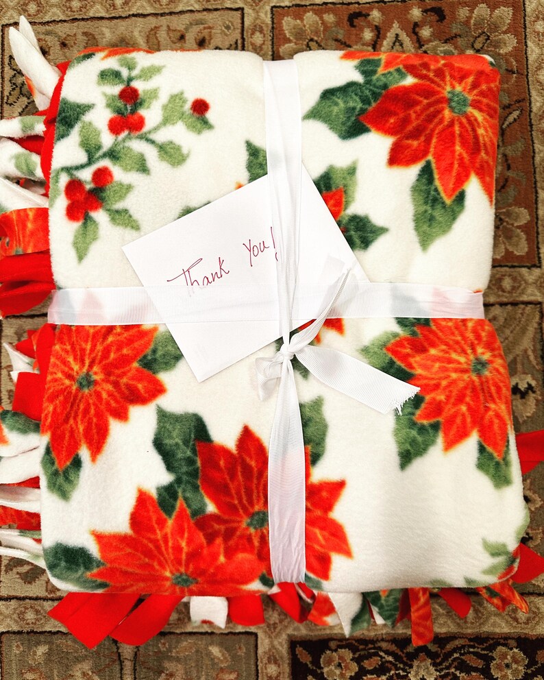 K&ouml;nnte beinhalten: Eine wei&szlig;e Fleece-Decke mit einem roten Weihnachtsstern-Druck und gr&uuml;nen Bl&auml;ttern. Die Decke ist mit einem wei&szlig;en Band gebunden und hat einen handgeschriebenen Zettel mit der Aufschrift "Danke!"