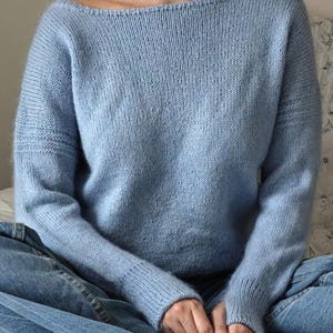 Peut inclure: Un pull en tricot bleu clair avec un col bateau et des manches longues. Le pull est fait d'un fil doux et texturé. La personne porte un jean bleu. Le pull est un vêtement confortable et élégant.