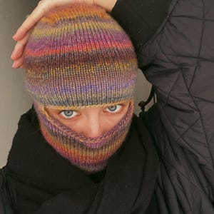 Peut inclure: Une cagoule en tricot rayée colorée avec un schéma de couleurs violet, orange et marron. La cagoule couvre la tête et le visage, seuls les yeux sont visibles.
