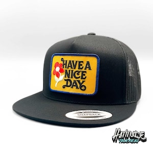 May include: Black trucker hat with a yellow patch that says "Have a Nice Day" with a red and white flower graphic. The hat has a black mesh back.
