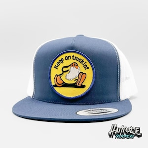 Op de afbeelding: Een blauwe en witte truckercap met een geel en blauw patch waarop "Keep on truckin'" staat en een cartoonfiguur.
