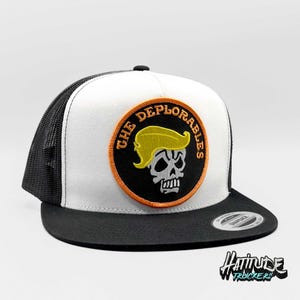 Puede incluir: Gorra de camionero blanca y negra con un parche bordado en negro y naranja que dice "The Deplorables" con una calavera con una peluca amarilla.