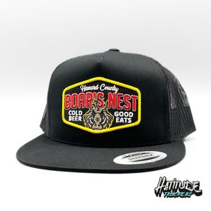 Könnte beinhalten: Schwarze Trucker-Kappe mit schwarzem Netzrücken. Die Vorderseite der Kappe zeigt einen roten und gelben gestickten Aufnäher mit der Aufschrift "Hazard County Boar's Nest Cold Beer Good Eats".