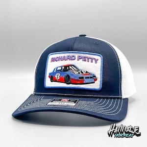 Puede incluir: Un gorro de camionero azul marino y blanco con un parche bordado que presenta el coche de carreras #43 de Richard Petty. El parche tiene un fondo blanco con acentos azules y rojos. El gorro tiene una parte posterior de malla blanca y una parte delantera azul marino.