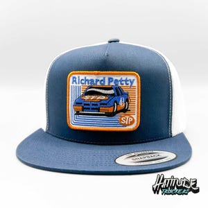 May include: A blue and white trucker hat featuring a patch with the text "Richard Petty" and a blue race car with the STP logo. The hat has a curved brim and a snapback closure.