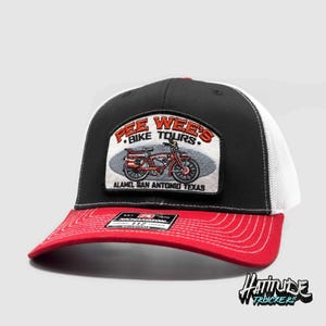 May include: A black and white trucker hat with a red peak. The hat features a patch on the front that reads "Pee Wee's Bike Tours" with a red bicycle illustration and the text "Alamo San Antonio Texas".